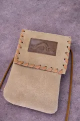 Cartera tipo bandolera de gamuza color arena, con solapa y detalles de costura en cuero marrón claro. Incluye una etiqueta de cuero grabada con el logo de la marca.
