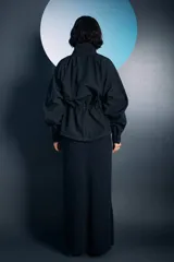 Campera negra de cuello alto con cierre frontal de botones a presión, mangas largas y cordones ajustables en la cintura. Presenta costuras a la vista en color blanco y un bordado decorativo en el pecho.