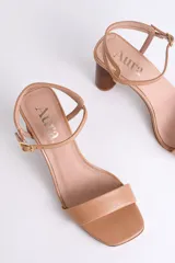 Sandalia de cuero color peltre, con tira ancha sobre los dedos y tira fina con hebilla al tobillo. El taco es cilíndrico de madera clara.