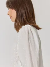 Camisa blanca de algodón con mangas largas abullonadas, detalle de volados en los hombros y puños elásticos. Presenta un diseño con cuello redondo y corte holgado.