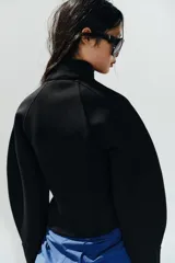 Chaqueta negra de cuello subido con cierre frontal de cremallera termosellada, manga larga con puños anchos y cremalleras laterales en los puños.