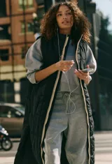 Chaleco largo acolchado de color negro, con cierre frontal completo y diseño sin mangas. Presenta un acabado matelasse y un borde contrastante en color claro a lo largo de la cremallera.