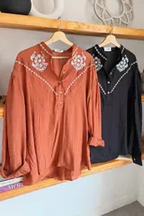 Camisa de color terracota con cuello camisero, cierre frontal de botones y mangas largas abullonadas. Presenta un delicado bordado floral en color blanco en la zona del pecho y los hombros, con un diseño de corte holgado.