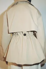 Trench corta de color beige con cuello de solapa y manga larga. Cuenta con bolsillos delanteros, cintura ajustable con cordones, bajo abullonado y cierre frontal cruzado con un botón visible. El interior está forrado con tela a cuadros.