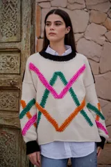 Sweater de punto color crudo con cuello redondo negro y bordados decorativos en forma geométrica en colores fucsia, verde y naranja. Tiene mangas largas con puños negros.