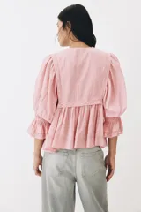Blusa rosada de algodón con ribetes de encaje, cuello redondo con abertura en V y tiras para anudar, mangas largas abullonadas con puños elásticos y terminación peplum con vuelo.