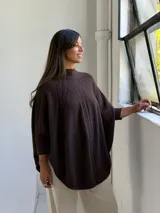 Poncho de punto color chocolate con cuello alto y diseño de trenzas verticales en el frente. Presenta mangas tres cuartos con puños acanalados y un corte holgado y fluido.