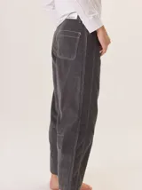 Pantalón de pana color gris oscuro, de corte barrel (holgado en caderas y muslos, ligeramente afín hacia el tobillo). Presenta costuras visibles en contraste de color amarillo claro, especialmente en la zona de la rodilla y el ruedo.