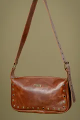 Cartera de cuero color marrón con diseño rectangular, cierre superior y detalles de tachas metálicas en todo el contorno. Incluye correa larga ajustable con hebilla.