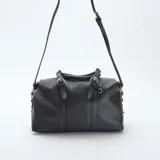 Bolso de mano estilo bowling en color negro, confeccionado en material texturizado. Presenta doble asa superior con detalles de hebillas decorativas, cierre principal con cremallera y correa larga ajustable y extraíble para llevar al hombro o cruzado. Incluye herrajes metálicos en tono dorado.