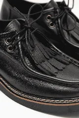 Mocasín de cuero negro con cordones y detalle de flecos en la capellada, con suela plana y costuras visibles en el borde.