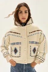 Campera de lana merino hervida en color crudo, con diseño de cuello camisero, cierre frontal de botones y bolsillos frontales. Presenta bordados artesanales con motivos geométricos en tonos marrón y azul en el frente, espalda y mangas.