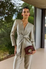 Blazer corto de satén color beige dorado, con escote en V profundo, hombreras y un lazo grande integrado en la cintura para anudar al frente. Presenta mangas largas de corte recto.