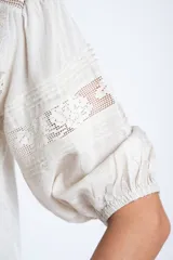 Blusa color crudo de manga corta abullonada, con escote en V y detalles de encaje y bordados verticales en el frente y mangas.