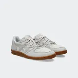 Championes Asics modelo Skyhand OG, con diseño retro inspirado en el balonmano de los años 90. Presenta una parte superior que combina gamuza suave y tejido texturizado en tonos gris claro, con detalles en color rosado. Incluye cordones estampados, un adorno tipo perla y una suela de goma color caramelo con amortiguación de EVA en el talón.