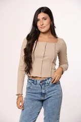 Remera de manga larga en color beige, confeccionada en tejido acanalado. Presenta un escote cuadrado y una abertura frontal con cierre de botones a presión. El diseño incluye costuras verticales decorativas y un ruedo con terminación irregular.