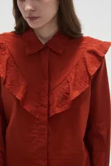 Camisa de algodón blanca con cuello camisero, cierre frontal de botones y volados bordados en el frente y hombros. Presenta mangas largas con puños acampanados y detalles bordados en el ruedo.