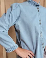 Camisa de denim con cuello alto tipo mao con volados, cierre frontal con botones y mangas largas con puños elásticos con volados.