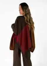 Ruana de tejido de punto con diseño de bloques de color en tonos tierra, incluyendo marrón, mostaza y crudo. Presenta terminaciones acanaladas en los bordes.