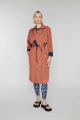 Trench largo cruzado color ladrillo, sin forro, con cinturón reversible azul marino y detalles de color de contraste ocultos. Tiene botones en el frente, bolsillos de vivo oblicuos y capucha oculta.