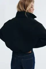 Campera negra de tejido efecto neopreno, con cuello chimenea alto, cierre frontal metálico y bajo fruncido con elástico. Presenta hombros caídos y corte corto.