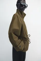 Parka amplia de Zara Woman Collection confeccionada en hilatura con mezcla de algodón, en color verde militar. Presenta cuello alto, manga larga con trabillas y botón, bolsillos laterales ocultos en costura, cintura ajustable con cordones y cierre frontal con botones.