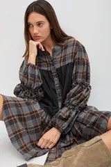 Vestido midi de corte holgado con diseño combinado en tela escocesa gris y naranja y panel central negro. Presenta cuello mao, cierre frontal con botones, mangas largas abullonadas con detalles de volados y puños elásticos.