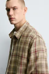Camisa de corte cropped confeccionada en algodón, con estampado de cuadros en tonos beige, marrón y rojo. Presenta cuello solapa, cierre frontal de botones, manga larga con puño abotonado y detalles de roturas distribuidas por toda la prenda con un bajo irregular.