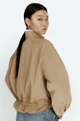 Campera oversized de sarga de algodón color beige, con cuello elevado, cierre frontal oculto y solapa rompevientos. Cuenta con mangas raglán, bolsillos delanteros con solapa y botón, y cordón ajustable en el bajo que frunce la cintura.