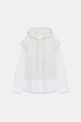 Camisa blanca de manga larga con un top de sudadera gris claro superpuesto, que incluye capucha ajustable con cordones. El bajo de la camisa tiene aberturas laterales.