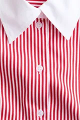 Camisa oversized de popelina de algodón con diseño a rayas verticales rojas y blancas. Presenta cuello clásico y puños anchos en color blanco sólido, cierre frontal con botones y bajo redondeado con aberturas laterales.