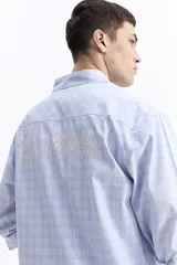 Camisa de popelina a cuadros, color celeste y blanco, de manga larga y corte holgado. Presenta un bolsillo frontal y cartera abotonada.