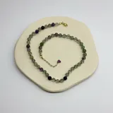 Collar de cuentas esféricas de piedra fluorita en tonos verdes y violetas, con separadores de nudos de hilo violeta. Incluye cadena de alargue y cierre con baño de oro.