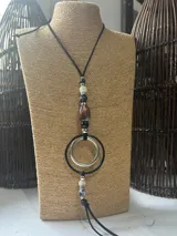 Colgante largo con cordón negro, decorado con una serie de cuentas de piedra en tonos blanco, negro y marrón, y un dije central compuesto por dos aros entrelazados, uno en color plata y otro en negro.