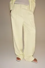 Pantalón de tiro medio de pierna ancha color amarillo pastel, confeccionado con mezcla de lino. Cuenta con bolsillos laterales ocultos, pliegues delanteros y cierre frontal con cremallera, botón y ganchos metálicos.
