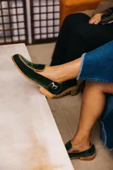 Mocasines de terciopelo verde oscuro con ribete tostado y detalle de aplique metálico plateado en forma de caballo en el talón. Cuenta con taco bajo y suela de madera.