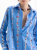 Camisa de calce holgado color azul, confeccionada en poplin satinado con estampado de motivos florales verticales. Presenta cuello clásico, mangas largas con puños abotonados y cierre frontal con botones.