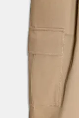 Pantalón de tiro alto estilo paperbag en color beige, con pinzas frontales y cinturón de la misma tela. Presenta bolsillos laterales tipo cargo con solapa y pernera ajustable con botones en el bajo.