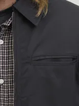 Campera estilo worker de color negro, con cuello camisero, cierre frontal con cremallera y bolsillos laterales. Presenta un diseño minimalista y funcional con acabado limpio.