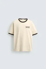 Camiseta de corte regular, color beige con cuello y bordes de mangas en color marrón oscuro. Presenta un diseño de texto bordado en el pecho con la frase 'Letting Time Pass By'.