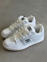 Championes DC modelo Manteca, color blanco con detalles en gris, cordones anchos y suela de goma marrón. Presentan el logo de DC en la lengüeta y los laterales.
