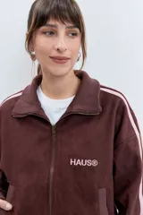 Cazadora de estilo deportivo en color marrón, confeccionada en tejido tipo gamuza. Presenta cierre frontal completo, cuello alto, puños y ruedo acanalados, y franjas laterales en tono rosado. Incluye el logo de la marca bordado en el pecho.