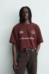 Camiseta de manga corta y cuello redondo, color granate oscuro, con estampado frontal en blanco que incluye texto y un escudo circular.