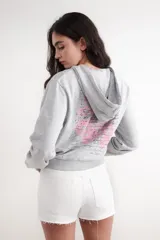 Buzo corto de felpa color gris melange con capucha, cierre frontal de cremallera y bolsillos tipo canguro. Presenta un estampado en tono rosado en el pecho.