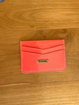 Tarjetero de cuero color coral con acabado brillante, diseño de ranuras en V y placa metálica con el logo de la marca en el frente.