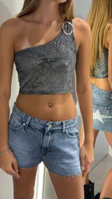 Top corto ajustado de una sola manga (asimetrico), color plateado con textura brillante o glitter. La única manga tiene un detalle de hebilla o anillo metálico en el hombro.
