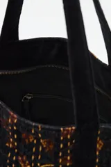 Bolso tote de algodón negro con estampado en tonos marrones y ocres, con costuras visibles en color claro. Presenta tiras de hombro negras y un adorno colgante con borlas negras y detalles dorados.