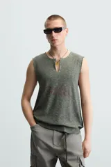 Camiseta sin mangas de punto acanalado, color verde oliva desgastado, con cuello redondo y abertura frontal en forma de 'V'. Presenta un estampado desvanecido en el centro del pecho.