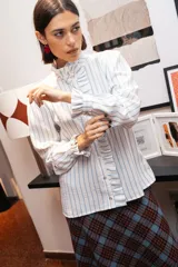 Camisa de manga larga con estampado de rayas verticales celestes sobre fondo blanco. Presenta un cuello alto con volados, cierre frontal con botones y puños con volados.
