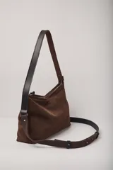 Cartera de hombro confeccionada en gamuza color chocolate, con cierre superior y doble sistema de correas: una fija de cuero liso negro para llevar al hombro y una correa larga ajustable y extraíble de gamuza y cuero negro para usar cruzada.